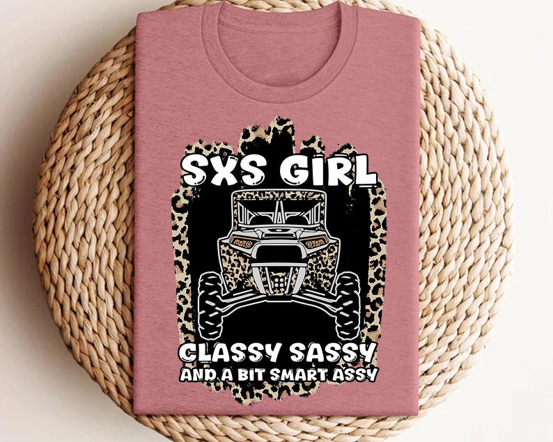 May include: A mauve crew neck t-shirt featuring a graphic of a side-by-side off-road vehicle over a leopard print background. The text reads SXS GIRL CLASSY SASSY AND A BIT SMART ASSY in white lettering with black outlines centered on the front.