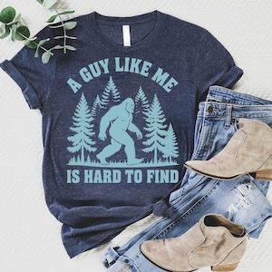 May include: Navy blue t-shirt with a Bigfoot graphic and the text "A GUY LIKE ME IS HARD TO FIND" in light blue. The shirt is paired with blue jeans and tan ankle boots. A casual, outdoorsy theme.