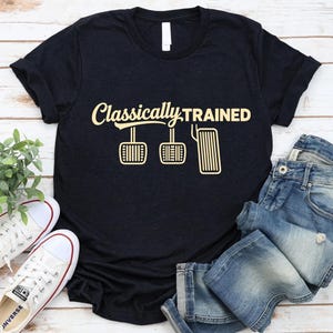 May include: Black t-shirt with the text "Classically Trained" in a cursive font, along with a graphic of car pedals. The shirt is paired with blue jeans and white sneakers with red accents.