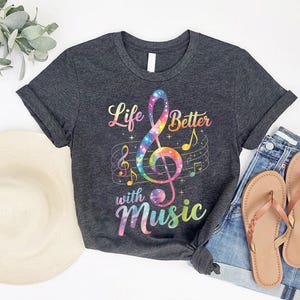 May include: Dark gray t-shirt with a colorful treble clef design and the words "Life Better with Music." The graphic includes musical notes and a rainbow color scheme. The shirt is paired with denim shorts and sandals.