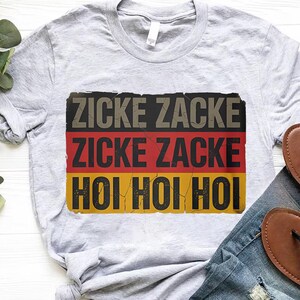 A gray t-shirt with a graphic design featuring the German text "ZICKE ZACKE ZICKE ZACKE HÖO HÖO" in red, black, and yellow colors.