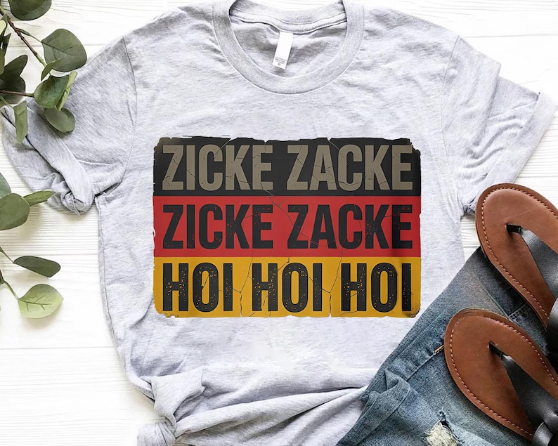 A gray t-shirt with a graphic design featuring the German text "ZICKE ZACKE ZICKE ZACKE HÖO HÖO" in red, black, and yellow colors.