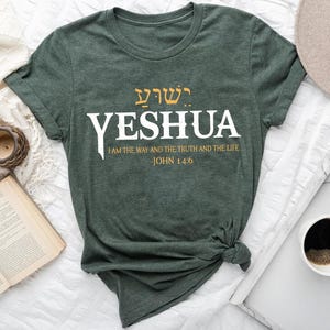 May include: A dark green t-shirt with the word "YESHUA" in large white letters. Above the word is Hebrew text in gold. Below the word is the text "I AM THE WAY AND THE TRUTH AND THE LIFE JOHN 14:6".