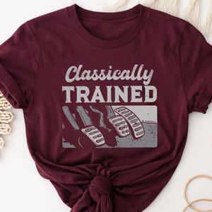 May include: A maroon short-sleeve t-shirt featuring the white text Classically Trained above a graphic illustration of three car pedals. The design shows a clutch, brake, and accelerator pedal in a stylized, textured white and grey print.