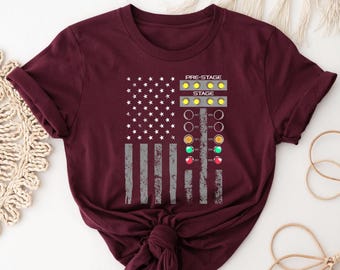 May include: A maroon short-sleeve t-shirt featuring a graphic of a distressed American flag combined with a drag racing Christmas tree light display. The graphic includes the text PRE-STAGE and STAGE above rows of yellow, green, and red lights.