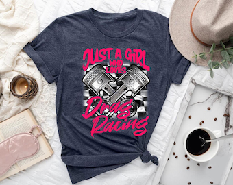 May include: A dark heather navy blue short-sleeve t-shirt featuring a graphic of two crossed engine pistons over a black and white checkered flag pattern. The text reads JUST A GIRL WHO LOVES DRAG RACING in a bold, bright pink stylized font.