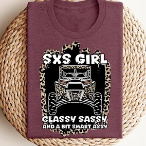 May include: A maroon t-shirt featuring a graphic print of a side-by-side vehicle centered on a leopard print background. The text reads SXS GIRL, CLASSY SASSY AND A BIT SMART ASSY in white lettering. The design is positioned on the front of the shirt.