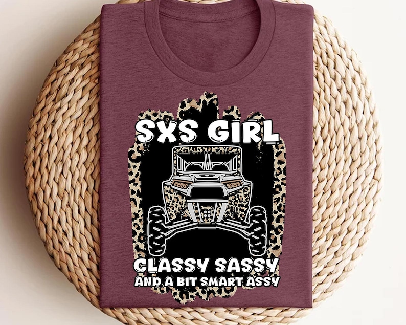 May include: A maroon t-shirt featuring a graphic print of a side-by-side vehicle centered on a leopard print background. The text reads SXS GIRL, CLASSY SASSY AND A BIT SMART ASSY in white lettering. The design is positioned on the front of the shirt.