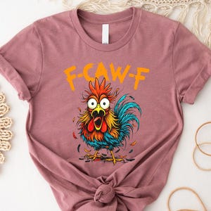 A pink t-shirt with a colorful illustration of a rooster and the text "F-CAWF-F" printed on it.