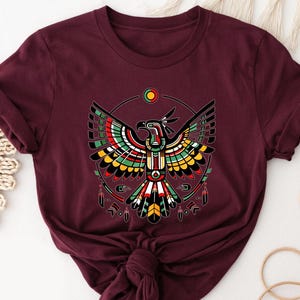 May include: A maroon short-sleeved t-shirt featuring a graphic print of a stylized eagle with outstretched wings. The bird is rendered in geometric patterns of red, yellow, green, and black, framed by a circular design with small hanging feathers.