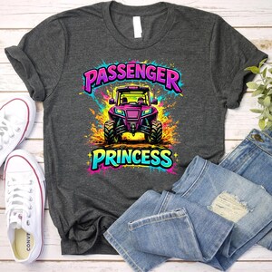 May include: Dark gray t-shirt with a colorful graphic of a purple off-road vehicle and the words "PASSENGER PRINCESS". The design has a neon color scheme. White canvas sneakers and blue jeans are also in the image.