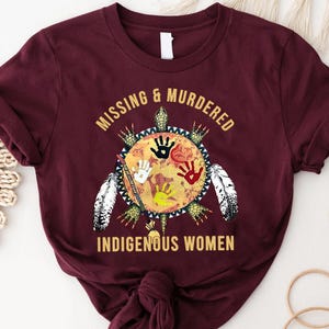 May include: A maroon short-sleeved t-shirt featuring a graphic of a turtle shell with four handprints in black, white, red, and yellow. The text reads MISSING & MURDERED INDIGENOUS WOMEN. The design includes feathers and a staff element.