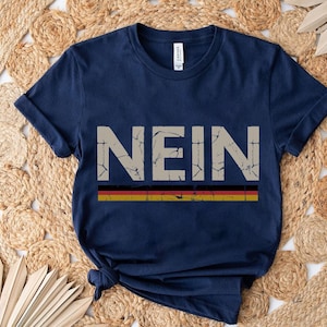 May include: Navy blue t-shirt with the German word "NEIN" in a distressed, off-white font. Below the word is a horizontal stripe in the colors of the German flag: black, red, and gold. The shirt is folded.
