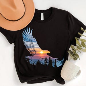 a black t-shirt with a graphic design depicting a soaring eagle against a sunset landscape, accompanied by a beige hat, a white mug, and a green plant.