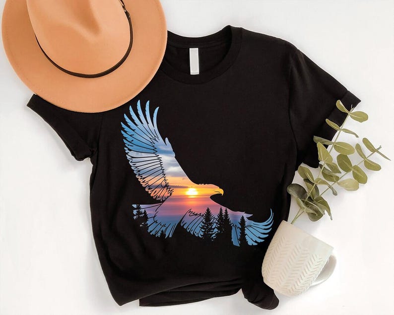 a black t-shirt with a graphic design depicting a soaring eagle against a sunset landscape, accompanied by a beige hat, a white mug, and a green plant.