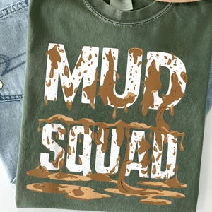 May include: Green t-shirt with the words "MUD SQUAD" in white block letters, with brown mud-like designs dripping from the letters. The shirt is displayed on top of blue jeans.