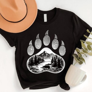 May include: Black t-shirt featuring a bear paw print design. The paw print includes a mountain landscape with a bear silhouette. A tan hat is visible in the upper left corner. The shirt is made of a soft material.