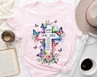 May include: Light pink t-shirt featuring a watercolor cross design with butterflies and floral accents. The cross has the text "GOD SAYS YOU ARE" with additional words like "UNIQUE" and "CHOSEN". The design includes colorful butterflies and flowers.