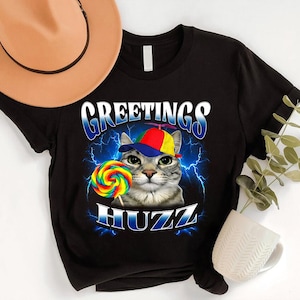 May include: Black t-shirt featuring a cat wearing a colorful hat and holding a rainbow lollipop. The shirt has the words "GREETINGS HUZZ" in a blue, stylized font with lightning bolt accents. A fun, whimsical design.