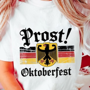 May include: White t-shirt featuring the words "Prost! Oktoberfest" in black script. The design includes a German flag and a black eagle emblem. The shirt is a casual wear item.