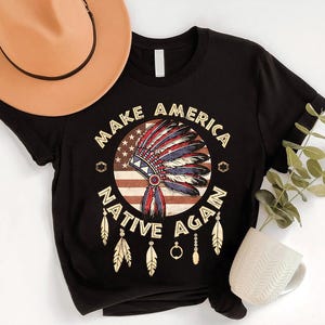 May include: Black t-shirt with a graphic design featuring a Native American headdress over an American flag. The text "MAKE AMERICA NATIVE AGAIN" is printed around the design in a circular pattern. The shirt is paired with a tan hat.