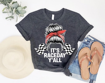 May include: Dark gray t-shirt with the text "IT'S RACEDAY Y'ALL" and a graphic of a woman with a red bandana and checkered flags. The shirt is paired with denim shorts and brown sandals.
