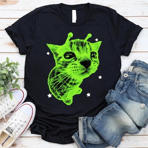 May include: Black t-shirt featuring a neon green alien cat graphic. The cat has large eyes and antennae, with white star accents. The shirt is paired with blue jeans and white sneakers on a light wood surface.