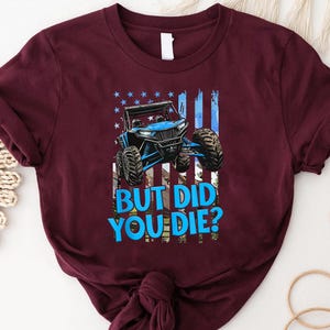 May include: Burgundy t-shirt featuring a blue off-road vehicle design with the text "BUT DID YOU DIE?" against a backdrop of a stylized American flag. The shirt is made of a soft material.