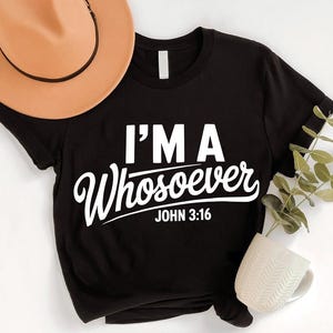 May include: Black t-shirt with the text "I'M A Whosoever JOHN 3:16" in white. A tan hat and a white mug are visible in the image. The shirt is a casual wear item.