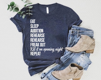 May include: Navy blue t-shirt with white text listing the steps of a performance: Eat, Sleep, Audition, Rehearse, Freak Out, Kill it on opening night, Repeat. The shirt is paired with blue jeans and tan ankle boots.