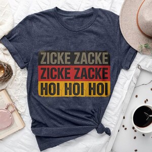 a blue t-shirt with the text "Zicke Zacke Hoi Hoi" printed on it, along with a hat, a book, a cup of coffee, and some other items arranged on a bed.