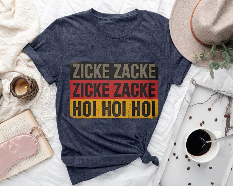 a blue t-shirt with the text "Zicke Zacke Hoi Hoi" printed on it, along with a hat, a book, a cup of coffee, and some other items arranged on a bed.