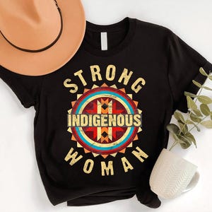 May include: Black t-shirt with a graphic design that reads "STRONG INDIGENOUS WOMAN" in a vintage style. The design features a circular emblem with geometric patterns in red, blue, yellow, and brown. A tan cowboy hat is visible in the upper left corner.