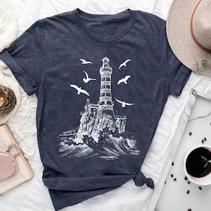 May include: Dark navy blue t-shirt featuring a white illustration of a lighthouse on a rocky island, with seagulls flying around it. The design includes crashing waves and a detailed depiction of the lighthouse structure. The shirt is knotted at the bottom.