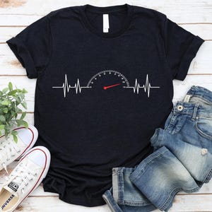 May include: Black t-shirt featuring a white speedometer design integrated with a heartbeat line. The speedometer needle is red. The shirt is paired with blue jeans and white sneakers.
