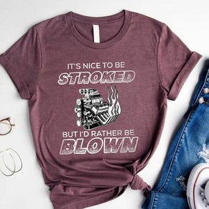 May include: A maroon short-sleeve t-shirt featuring a graphic of a car engine with flames and the white text IT'S NICE TO BE STROKED BUT I'D RATHER BE BLOWN. The shirt has a crew neckline and a standard fit, displayed flat with a casual aesthetic.