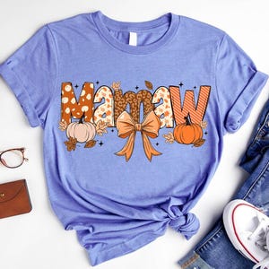May include: A light purple short-sleeve t-shirt featuring the text Mamaw in large, patterned letters. The design includes orange pumpkins, brown autumn leaves, and a central orange bow. The letters have leopard print and striped patterns.