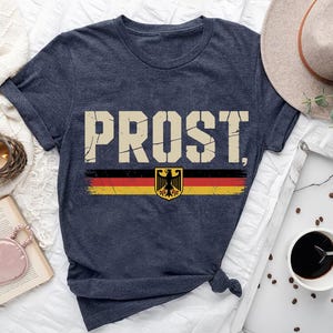 May include: Navy blue t-shirt with the word "PROST" in distressed, off-white lettering. Below the text is a horizontal stripe in the colors of the German flag, with a German coat of arms. The shirt is knotted at the bottom.