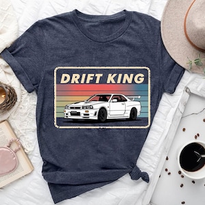 May include: Navy blue t-shirt featuring a white sports car graphic with the text "DRIFT KING" in a rectangular design. The design has a retro sunset color scheme. The shirt is made of a soft material.