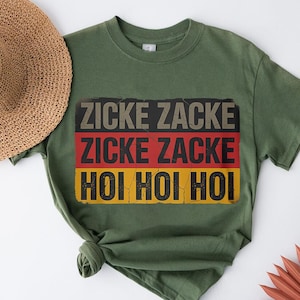 A green t-shirt with the text "ZICKE ZACKE ZICKE ZACKE HÖI HÖI" printed on it, along with a straw hat and some red objects in the background.