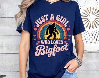 May include: Navy blue t-shirt with a colorful graphic. The graphic features a silhouette of Bigfoot in front of a rainbow, with the text "JUST A GIRL WHO LOVES Bigfoot" in pink and white.