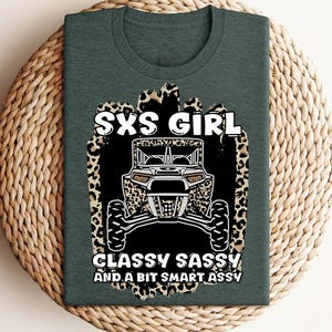 May include: A dark green t-shirt featuring a graphic print of an off-road vehicle centered on a leopard print background. The text reads SXS GIRL CLASSY SASSY AND A BIT SMART ASSY in white capital letters. The design is positioned on the front chest.