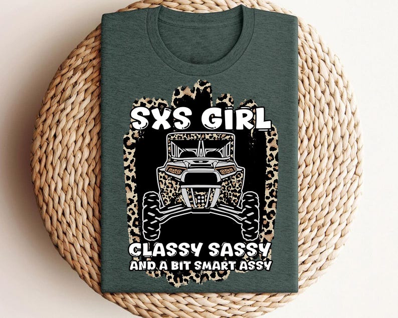 May include: A dark green t-shirt featuring a graphic print of an off-road vehicle centered on a leopard print background. The text reads SXS GIRL CLASSY SASSY AND A BIT SMART ASSY in white capital letters. The design is positioned on the front chest.
