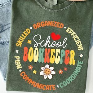 May include: A green t-shirt with the words "School Bookkeeper" in a colorful, retro font. The design includes a red heart, flowers, and words like "Organized" and "Efficient" around the central text. The shirt is displayed on a pair of blue jeans.