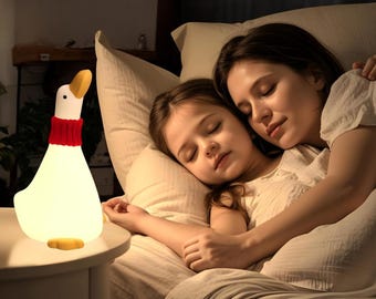 May include: A white goose-shaped nightlight with a red scarf sits on a bedside table next to a mother and child sleeping in bed.