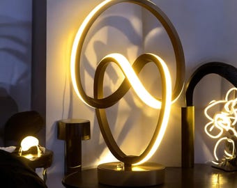 May include: A modern table lamp with a unique, abstract design. The lamp features a black metal base and a white LED light that illuminates the space with a warm glow.