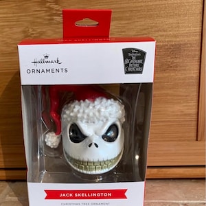 May include: A Jack Skellington Christmas tree ornament. The ornament features Jack Skellington's skull-like face wearing a red Santa hat with white trim. The ornament is in a clear plastic box with the Hallmark Ornaments logo and "The Nightmare Before Christmas" branding.