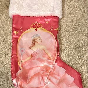 May include: Pink Christmas stocking with a white faux fur cuff. The stocking features a princess illustration in a gold frame. The princess is wearing a pink gown and holding a wand. The stocking is made of a shiny, satin-like material.