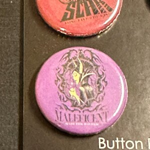K&ouml;nnte beinhalten: Ein Satz runder Kn&ouml;pfe mit Charakterdesigns. Ein Knopf ist lila mit dem Wort "Maleficent" und einer Charakterillustration. Ein weiterer Knopf ist rot mit dem Wort "Scar" und einer Charakterillustration.