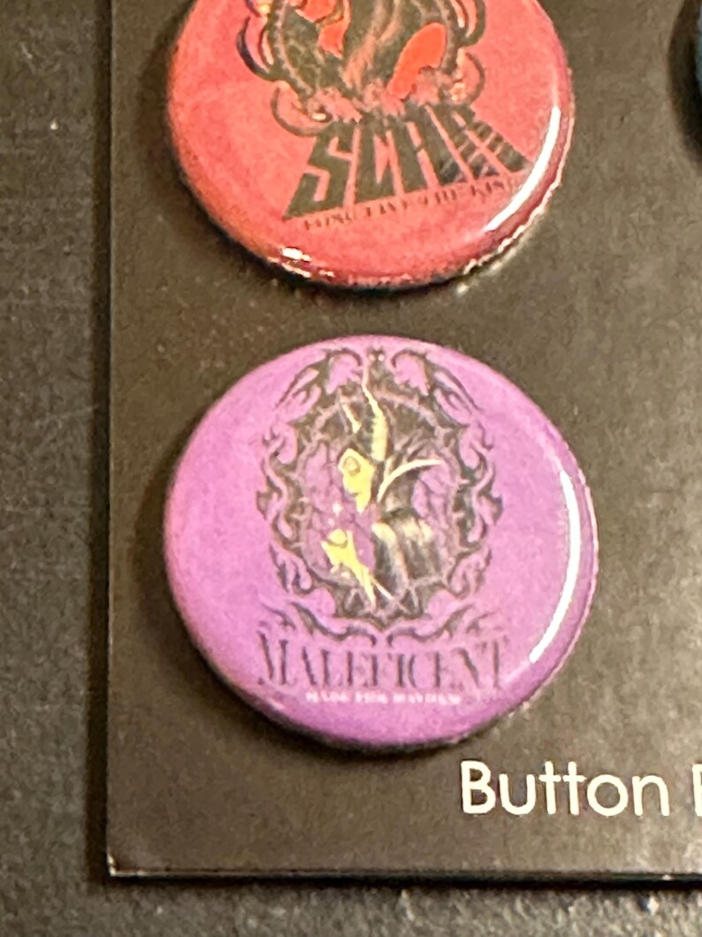 K&ouml;nnte beinhalten: Ein Satz runder Kn&ouml;pfe mit Charakterdesigns. Ein Knopf ist lila mit dem Wort "Maleficent" und einer Charakterillustration. Ein weiterer Knopf ist rot mit dem Wort "Scar" und einer Charakterillustration.